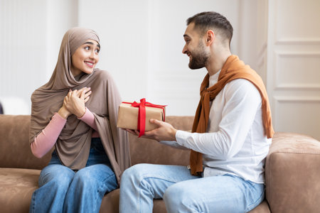 Mari musulman donnant une boîte-cadeau emballée à une femme surprise célébrant les vacances de noël ou la saint-valentin ensemble assis sur un canapé à la maison. concept de romance, de fêtes, de cadeaux et de cadeaux