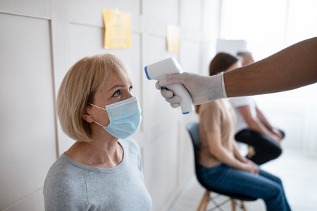 Senior woman having her temperature checked before coronavirus vaccine injection at hospital waiting room. contagious disease prevention, covid-19 immunization campaign Фото со стока