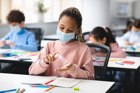 Hygiene and health concept. african american girl applying antibacterial hand sanitizer spray on palm, wearing medical face mask, sitting in classroom at school. prevent spreading of infection