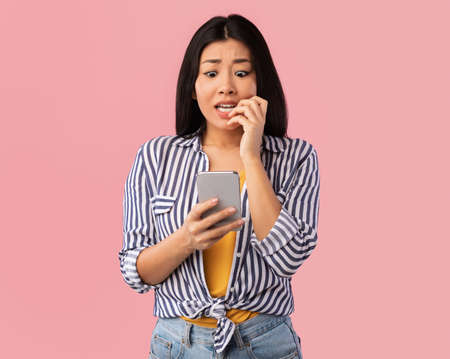 Portrait of anxious scared asian young woman looking at mobile phone seeing bad news, photos or message. worried lady biting nails isolated on pink studio wall background. human reaction, expression