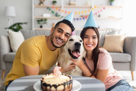 Happy multiracial couple celebrating their dogs birthday with festive cake, wearing party hats at home
