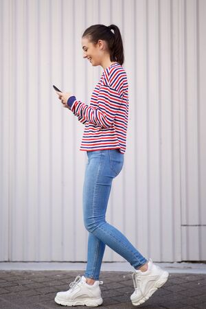 Full body side portrait of happy young woman walking with cellphone