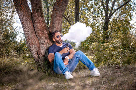 Stylish brutal vape man enjoying an e-cigarette in the woods. handsome man.
