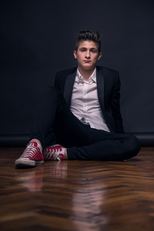 One young teenage boy black background studio formal clothes sneakers sitting Imagens