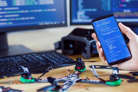 Closeup programmer hand is holding smartphone with program code software on screen for controlling fpv drone man developer is building constructing quadcopter from kit with microcontrollers