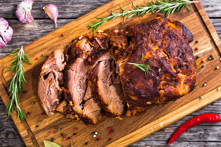 grilled pork: Oven-Barbecued Pork shoulder, cut on slices on chopping board with rosemary, peppercorn and bay leaf on old dark planks with garlic and chili on background, view from above, close-up