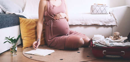 Pregnant Woman Packing Bag For Maternity Hospital, Making Notes, Checking List In Diary. Expectant Mother With Suitcase Of Baby Clothes And Necessities Preparing For Newborn Birth During Pregnancy.