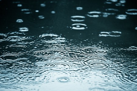 Rain drops rippling in a puddle with blue sky reflection Фото со стока