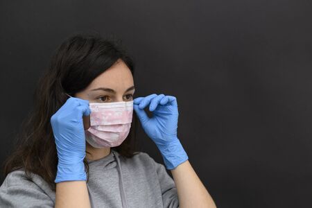 The young dark-haired girl with brown eyes puts on a one-time mask in blue gloves. he does not look at the camera, but to the side with a sad look. protective light mask and gloves, a medical kit, they are used in hospitals as part of a uniform by doctors of various specialties, such as surgeons or nurses. used in the professional beauty industry, cosmetology and beauty salons. also used by sick people. if the person is a carrier of a virus or infection, he is recommended to wear such a protective mask, otherwise he can infect other people. it is better for a person with a cold, cough, fever, flu to stay at home, to isolate himself or to quarantine. in an epidemic, pandemic or environmental pollution, it is safer to carry disposable masks, gloves and an antiseptic, that is, a disinfectant. hand and face hygiene is important. this is a way to reduce the spread of the virus. your safety is your health. in the midst of illness, there may be a shortage of this product, a crisis, because people are in a panic buying everything. composition on a black background, horizontal view.