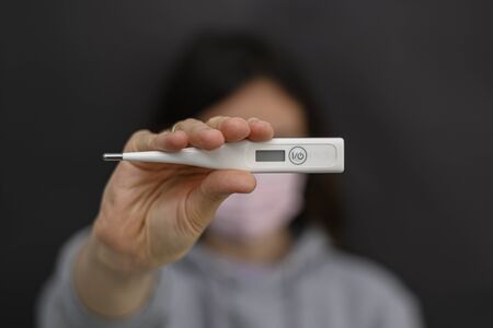 Self-monitoring and prevention during infections and pandemics. Temperature measurement is one of the important control points. The girl holds a thermometer in her right hand, there is no manicure. The thermometer is off, it does not show anything. He is in focus. The background where the girl stands is blurred. During outbreaks of viruses, infections, flu and coronovirus, bacterial sites should be treated. Medical protective equipment is used in hospitals by doctors of various specialties, such as surgeons or nurses. Used in the professional beauty industry, cosmetology and beauty salons. Also used by sick people. If the person is a carrier of a virus or infection, he is recommended to wear a protective mask, otherwise he can infect other people. It is better for a person with a cold, cough, fever, flu to stay at home, to isolate himself or to quarantine. In an epidemic, pandemic, or environmental pollution, it is safer to carry disposable masks, gloves, and an antiseptic, that is, a disinfectant. Hand and face hygiene is important. This is a way to reduce the spread of the virus. Your safety is your health. In the midst of illness, there may be a shortage of this product, a crisis, because people buy everything in a panic. Horizontal view. Close-up. Copy space. Dark background. - 142962434