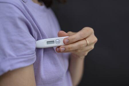 Self-monitoring and prevention during infections and pandemics. temperature measurement is one of the important control points. the girl is going to measure the temperature, holding a thermometer under the armpit. no manicure. the thermometer is still off, it does not show anything. he is in focus. the background where the girl stands is blurred. during outbreaks of viruses, infections, flu and coronovirus, bacterial sites should be treated. medical protective equipment is used in hospitals by doctors of various specialties, such as surgeons or nurses. used in the professional beauty industry, cosmetology and beauty salons. also used by sick people. if the person is a carrier of a virus or infection, he is recommended to wear a protective mask, otherwise he can infect other people. it is better for a person with a cold, cough, fever, flu to stay at home, to isolate himself or to quarantine. in an epidemic, pandemic, or environmental pollution, it is safer to carry disposable masks, gloves, and an antiseptic, that is, a disinfectant. hand and face hygiene is important. this is a way to reduce the spread of the virus. your safety is your health. in the midst of illness, there may be a shortage of this product, a crisis, because people buy everything in a panic. horizontal view. close-up. copy space. dark background. Banco de Imagens