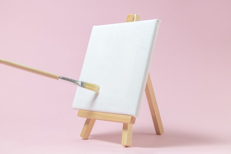 blank art canvas on easel and paintbrush against pastel pink stock