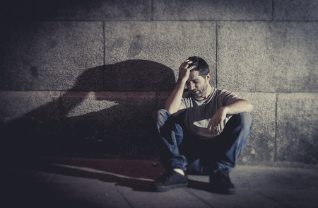 sad lonely: white wasted young man sitting on street ground with shadow on concrete wall feeling miserable and sad in urban scene representing depression and sickness