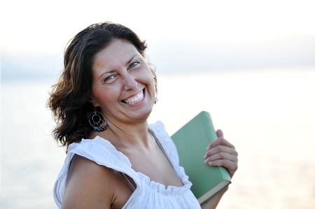穏やかでリラックスした夏の夜を楽しんで海の前にビーチの屋外ポートレートに笑みを浮かべて本を持って彼女の 40 代で幸せな魅力的な成熟した女性 の写真素材 画像素材 Image