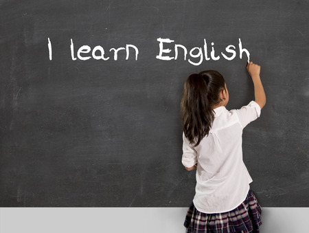 sweet little Hispanic girl at school lesson with ponytail and uniform writing with chalk on classroom blackboard in English learning , wisdom and successful education concept