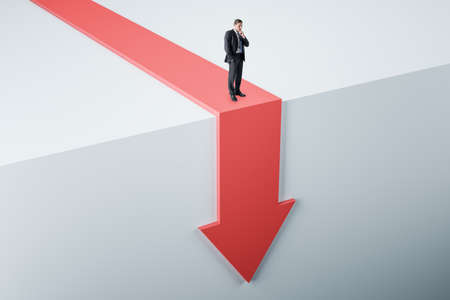 Personal crisis and business decline concept with confused businessman on red falling down arrow with abstract light backdrop Personal crisis and business decline concept with confused businessman on red falling down arrow with abstract light backdrop