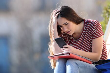 Worried student checking bad news on mobile phone sitting in a campus