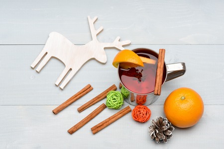 hot drink of mulled wine or tea with cinnamon spice stick in metallic mug near christmas decorative wood deer, ball, orange fruit and pinecone on grey wooden background
