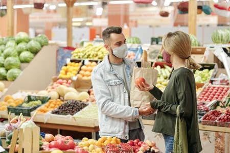 Young couple in cloth masks standing at counter and buying organic food at market: girl choosing tomatoes