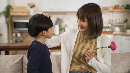 Garçon asiatique donnant un baiser sur le visage et la fleur d'oeillet à son mon le jour de la mère. maman souriante touchant la tête de son bébé et le louant sur un canapé à la maison
