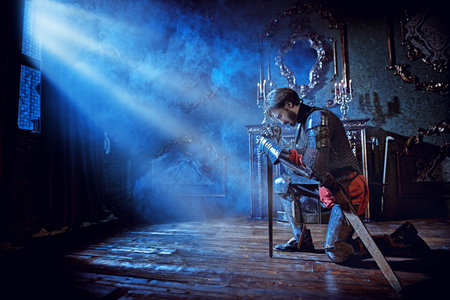 Medieval knight in armor makes an oath of allegiance by kneeling and bowing his head on the sword in the castle before the battle. Фото со стока