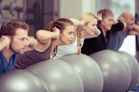 Group of diverse people in a receding line doing pilates in a gym balancing over the gym balls with their hands laocked behind their necks toning their muscles Фото со стока