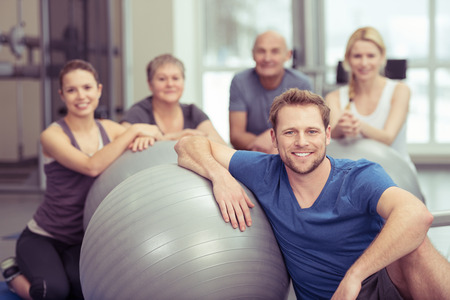 Smiling handsome young man in pilates class at the gym sitting leaning on his gym ball with the rest of the diverse group gathered behind him