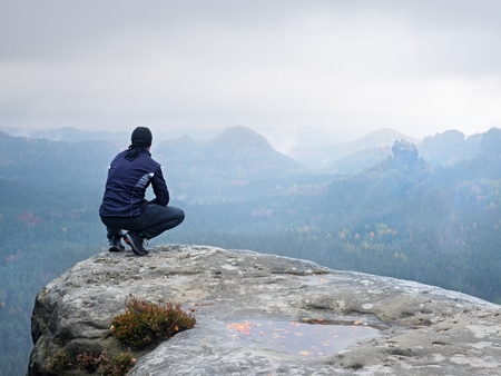 Adult Tourist In Black Trousers Jacket And Cap Sit On Cliffs Edge Looking To Misty Adult Tourist In Black Trousers Jacket And Cap Sit On Cliffs Edge Looking To Misty
