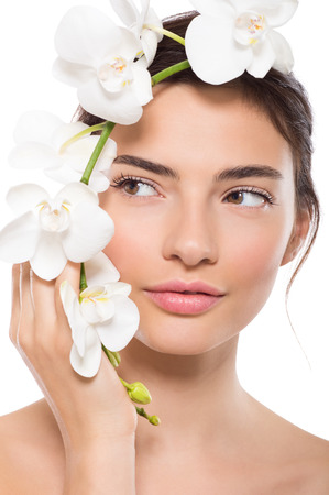 Closeup face of beautiful young woman with orchid flower looking away. Portrait of beauty girl holding flowers near face isolated on white background. Beauty treatment and skin care concept. Фото со стока