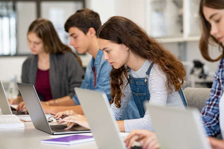 Serious college students studying on laptop sitting in a row in library. young university multiethnic students using computer for study in classroom. side view of casual girl typing on laptop during computer lesson. Фото со стока