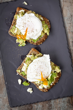 Smashed avocado and feta cheese toast with poached eggs. Overhead view, on dark slate. Фото со стока