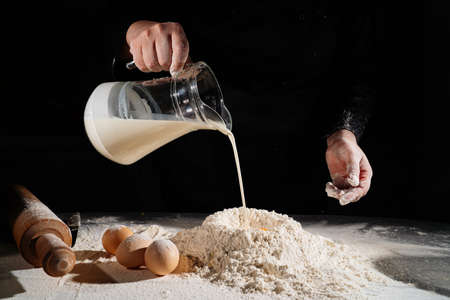 Une femme verse du lait d'une cruche dans de la farine pour pétrir la pâte sur fond noir.