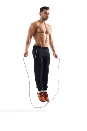 Side view of shirtless caucasian bodybuilder jumping in place using rope. muscular man with perfect tensed body looking straight, isolated on white studio background. concept of bodybuilding, fitness.