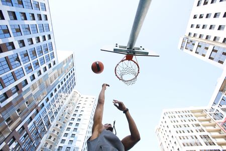 Low angle view at african basketball player shooting slam dunk against sky in urban background, copy space
