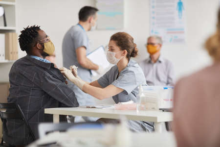 Side view portrait of young female doctor vaccinating african american man in medical clinic, copy space