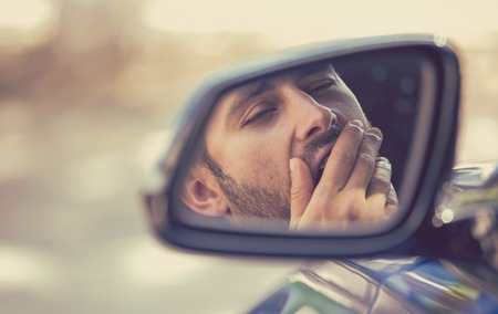Side mirror view reflection sleepy tired fatigued yawning exhausted young man driving his car in traffic after long hour drive. Transportation sleep deprivation accident concept