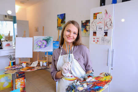 かわいい女性画家と壁の白い絵画スタジオに立って手でブラシとカラフル パレット笑顔 ポーズ 笑うとカメラの前で浮気します 女の子欧州外観大人と子供のための美術学校を宣伝します の写真素材 画像素材 Image