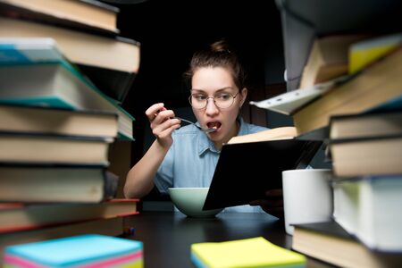 Girl student takes food while studying at home in the evening. on the table is a large number of books