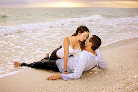 Two lovers on the beach, he is laying on his back, she is dripping wet between his legs making eye contact with him, they are wet from the ocean waves. In the background the sun is setting. Stock Photo