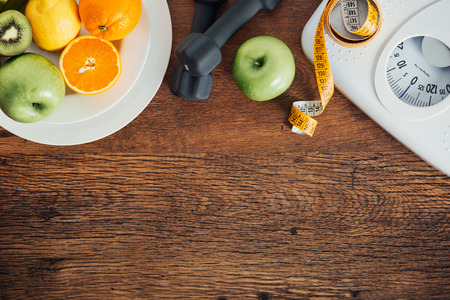 Concept de remise en forme et de perte de poids, haltères, échelle blanche, fruits et ruban à mesurer sur une table en bois, vue de dessus