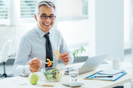 Smiling businessman sitting at office desk and having a lunch break, he is eating a salad bowl - 51616896