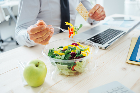Der Geschäftsmann, der eine Mittagspause am Schreibtisch hat, isst er frischen Salat und hält einen Cracker, unerkennbare Person