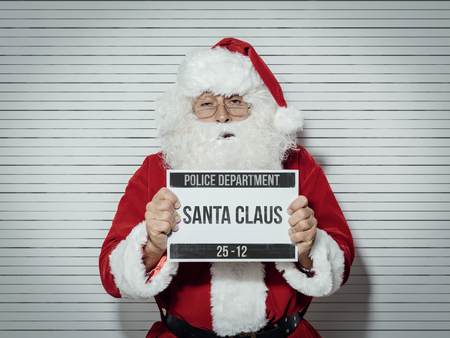 Santa claus arrested on christmas eve, he is posing for his mug shot at the police department and holding an identification board