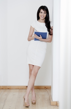 Young woman in white dress leaning against a white wall holding a blue folder Stockfoto