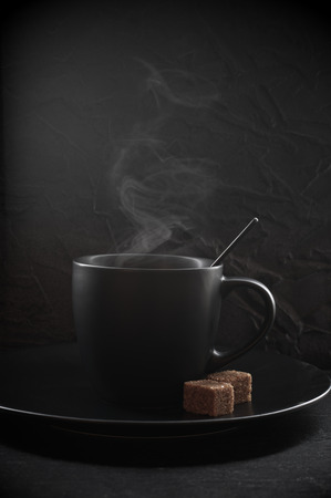 Black cup of hot coffee with steam and brown sugar cubes in plate on dark background.