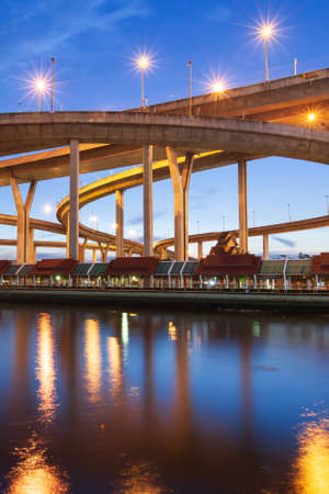 Picturesque low angle view of highway interchange and suspension bridges at twilight, glowing lights reflection on surface of chao phraya river, thailand. long exposure.