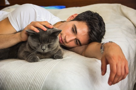 Handsome Young Animal-Lover Man on a Bed, Hugging and Cuddling his Gray Domestic Cat Pet.
