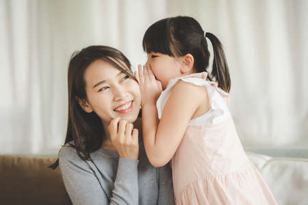 Little asian daughter girl whispering i love you mommy to happy smiling mother in living room Фото со стока Little asian daughter girl whispering i love you mommy to happy smiling mother in living room Фото со стока