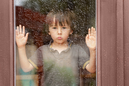 Little boy behind the window in the rain, looking sad Фото со стока