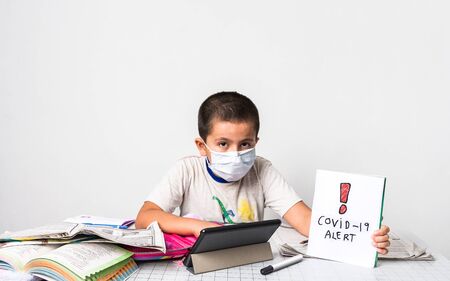 Corona virus / home schooling concept image with a young boy wearing a mask sitting at a table studying - health care and education concept image with copy space for text. Фото со стока